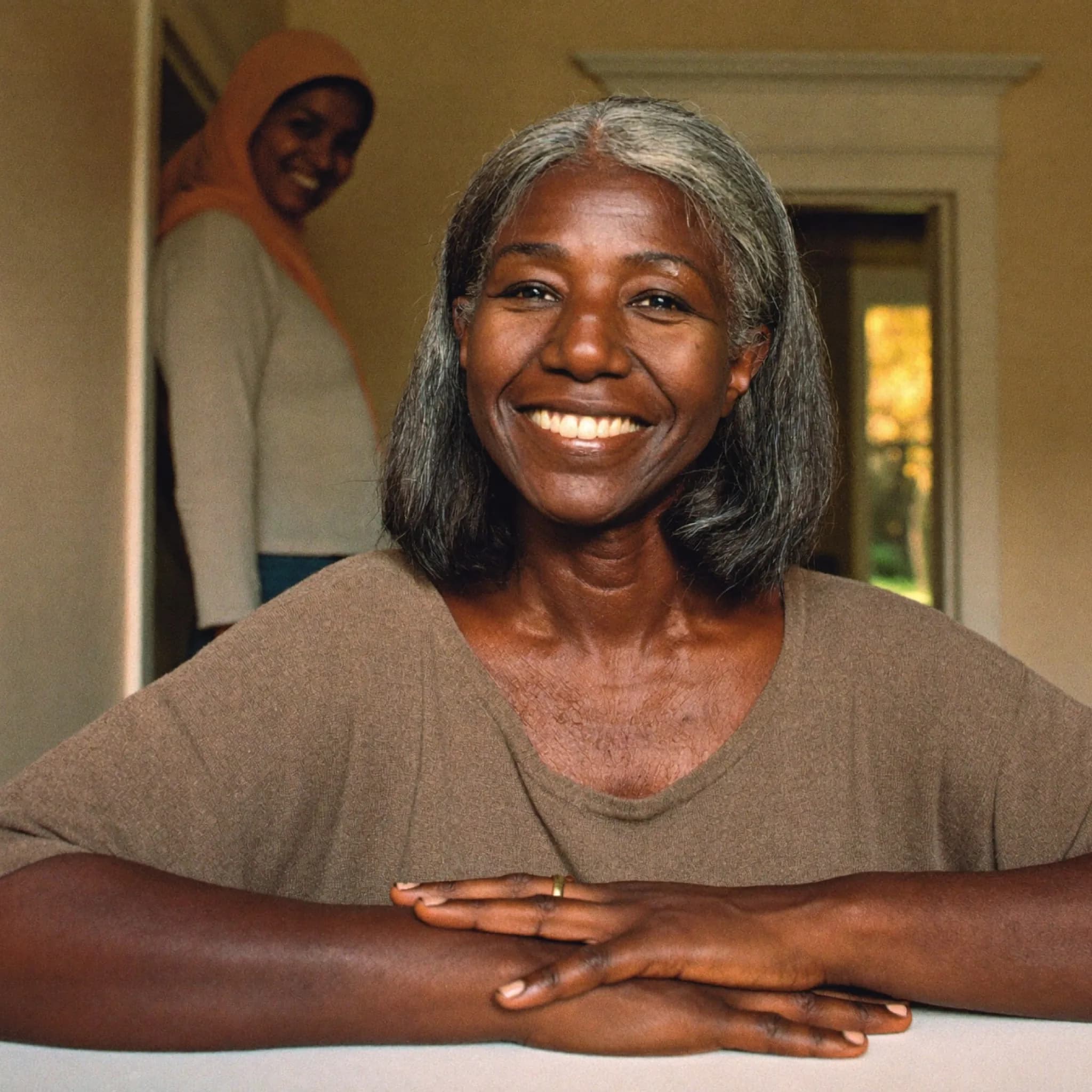 A woman smiling warmly towards the camera at a kitchen table, with a friend in the doorway behind her